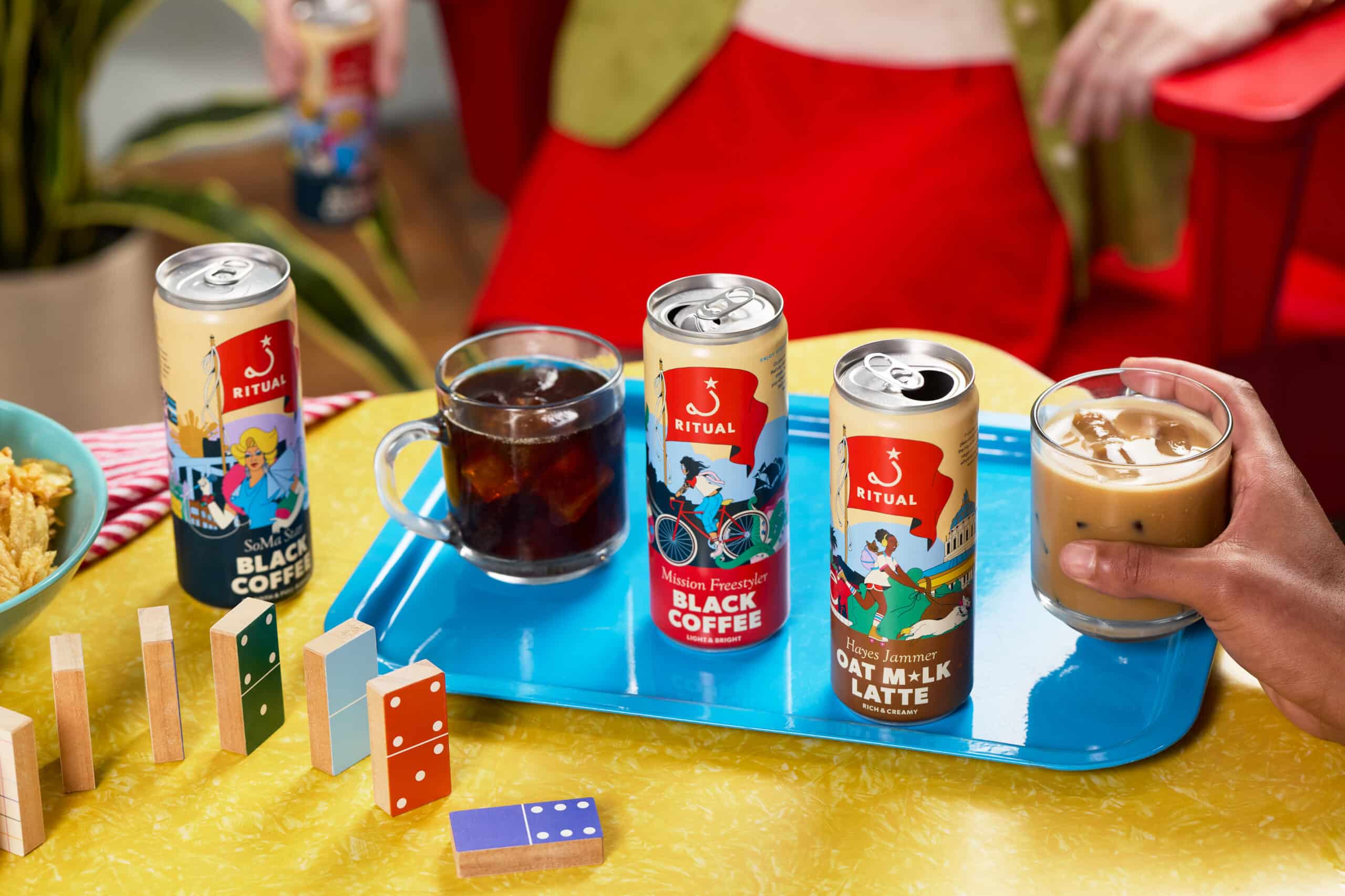 Cans of Ritual cold coffee on a yellow table surrounded by colorful dominos and a bowl of chips
